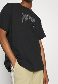 Černé bavlněné tričko se zkresleným bílým textovým grafickým prvkem s nápisem "NIGHT RIDER." Volný střih s krátkými rukávy a kulatým výstřihem.