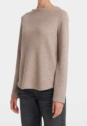 Haut beige à manches longues en maille gaufrée porté avec un jean gris foncé, montré du milieu du torse au cou et aux poignets sur un fond uni.
