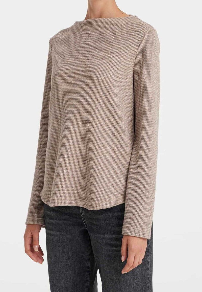 Haut beige à manches longues en maille gaufrée porté avec un jean gris foncé, montré du milieu du torse au cou et aux poignets sur un fond uni.