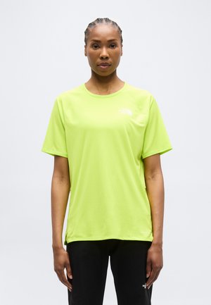 Kvinde med flettet hår iført en lysegrøn kortærmet atletik-T-shirt og sorte bukser, stående mod en ensfarvet baggrund.