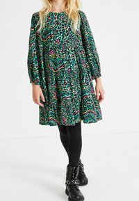 Zelené šaty s potiskem a růžovými a černými akcenty; dlouhé vycpané rukávy, nařasený pas a rozšířená sukně. Nosené s černými legínami a botami.