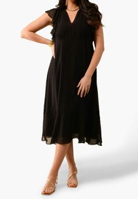 Musta pilkullinen midi-mekko, jossa on röyhelöhihoja, V-pääntie ja valuva helma. Vikattuna kultaisilla nauhaliinoilla ja korulla.