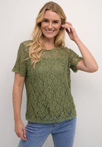 Top di pizzo verde con maniche corte, motivo floreale, scollatura e orlo scallop, indossato sopra jeans blu. La texture appare morbida e delicata.