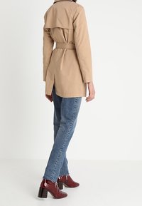 Trench-coat beige avec une taille ceinturée, un col classique et un détail dos superposé. Associé à un jean bleu et des bottines bordeaux.