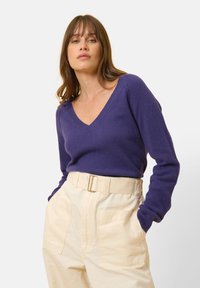 Fialový svetr s výstřihem do V, vyrobený z měkkého materiálu, spárovaný s krémovými kalhotami s vysokým pasem, které mají široký pásek a přední kapsy.