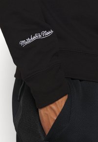 Felpa nera con polsini a coste, caratterizzata da un logo "Mitchell & Ness" ricamato bianco sulla manica. Indossata con pantaloni sportivi neri.