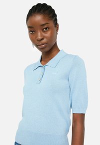Polo shirt tricoté bleu clair avec des manches courtes, des poignets côtelés et un col, arborant un petit logo brodé et une patte de boutonnage à trois boutons.