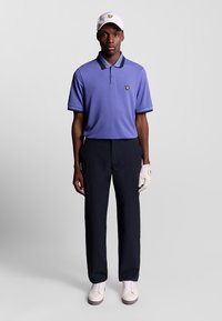 Uomo che indossa una polo viola, pantaloni neri, guanto da golf bianco, cappellino bianco e scarpe bianche, in piedi contro uno sfondo neutro.