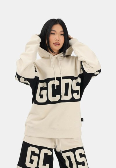 Nuori nainen säätää beige-mustan hupullisen hupparin huppua ja siihen sopivia shortsseja, joissa on suuri GCDS-logo edessä valkoista taustaa vasten.