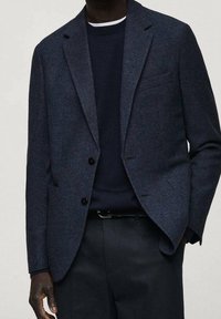 Granatowa marynarka o strukturze tkaniny, z dwoma guzikami, klapami z wycięciem oraz kieszenią na lewej piersi, zestawiona z ciemnym swetrem i spodniami.