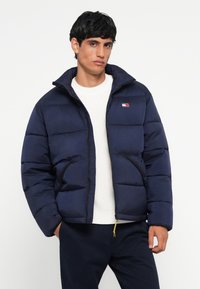 Marineblaue Steppjacke mit gepolsterten Bereichen, hohem Kragen, Reißverschluss und seitlichen Taschen. Mit kleinem Logo auf der linken Brust.