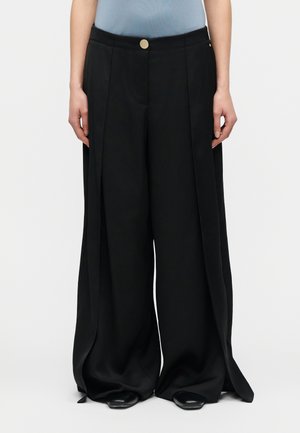 Personne portant un pantalon noir à jambes larges avec des plis avant et une fermeture à bouton doré, associé à des chaussures noires pointues et un haut bleu clair.