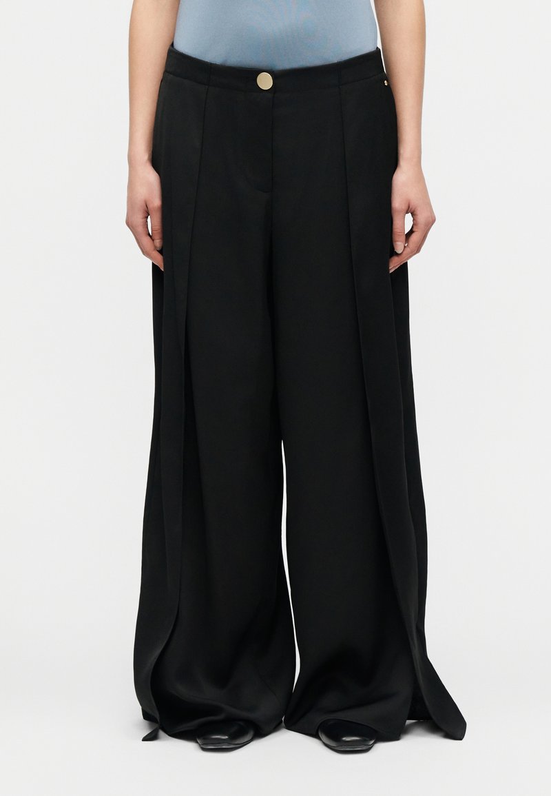 Άτομο με μαύρο παντελόνι φαρδιάς γραμμής με πιέτες μπροστά και κλεισίματα με χρυσό κουμπί, συνδυασμένο με μαύρα μυτερά παπούτσια και ανοιχτό μπλε τοπ.