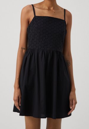 Kobieta w czarnej sukience bez rękawów z fakturowanym, haftowanym kwadratowym dekoltem i marszczoną spódnicą, stojąca na jednolitym tle.