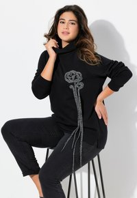Felpa nera con collo alto, decorata con un design floreale ricamato in argento e lunghi dettagli pendenti. Jeans neri aderenti completano l'outfit.