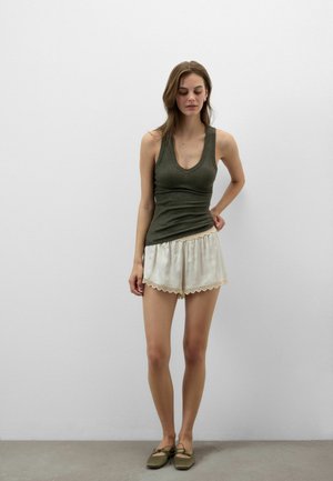 Vrouw staat tegen een witte muur, draagt een olijfgroene mouwloze top, crèmekleurige shorts met kanten rand en olijfgroene platte schoenen met strikjes.
