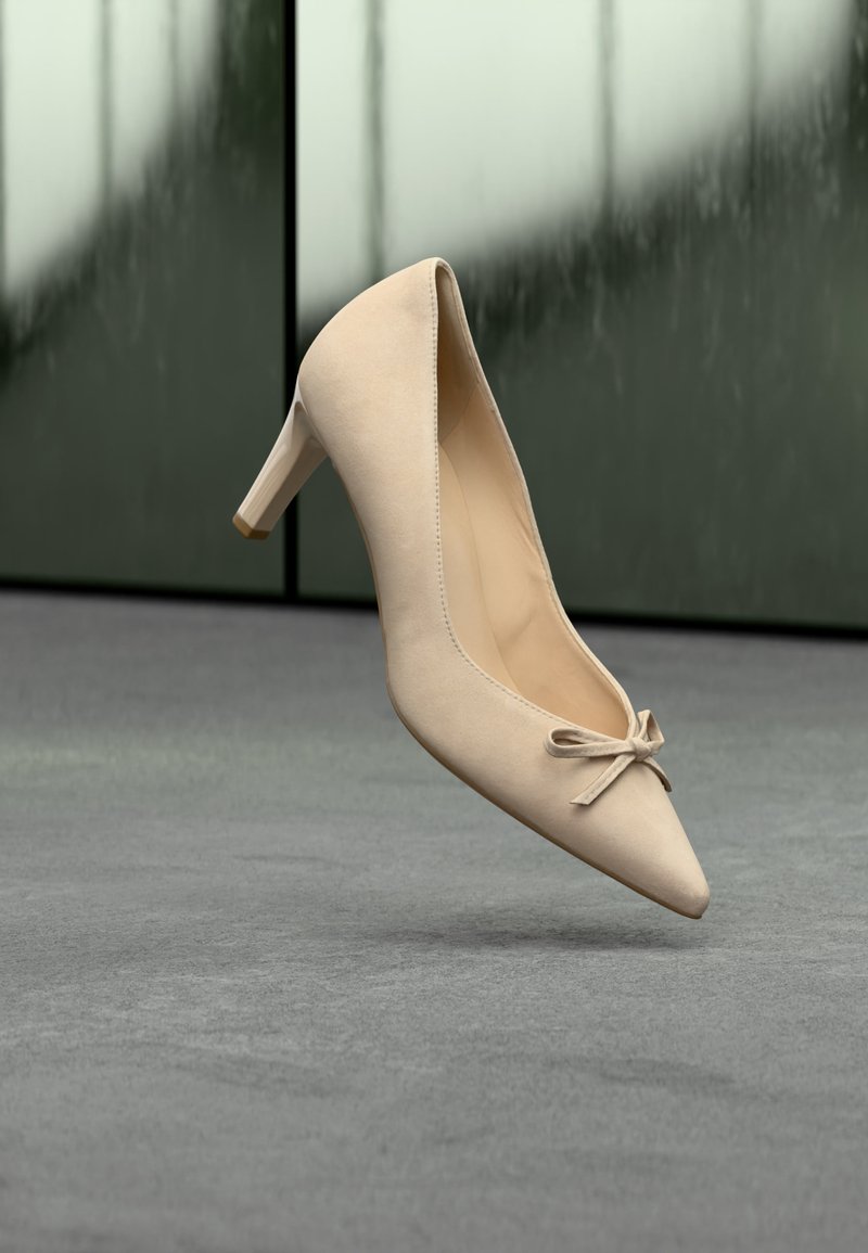 Μπεζ γόβα με ψηλό τακούνι, μυτερή μύτη και μικρό φιόγκο που αιωρείται πάνω από γκρι πάτωμα, με θολό γυάλινο φόντο.