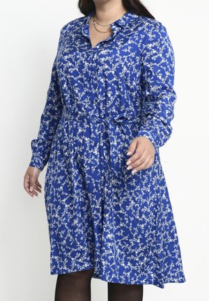 Femme portant une robe bleue à manches longues avec un motif floral blanc, un col et une ceinture nouée à la taille, associée à des collants noirs.