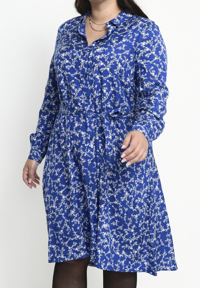 Femme portant une robe bleue à manches longues avec un motif floral blanc, un col et une ceinture nouée à la taille, associée à des collants noirs.