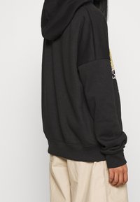 Svart huvtröja med känguruficka, ribbade ärmslut och avslappnad passform. Har färgglada grafiska accenter på baksidan.