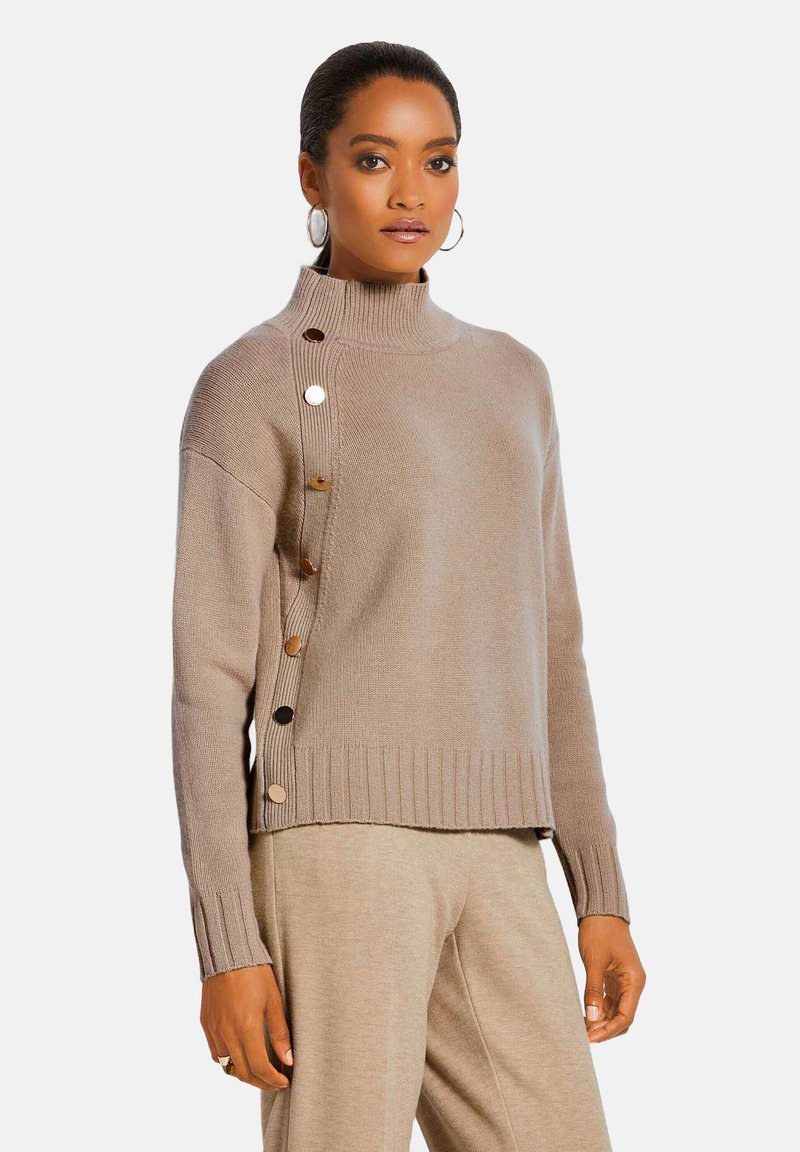 Beige Strickpullover mit hohem Kragen, geripptem Saum und seitlicher Knopfdetails. Weiche Textur und lockere Passform. Kombiniert mit beigen Hosen.