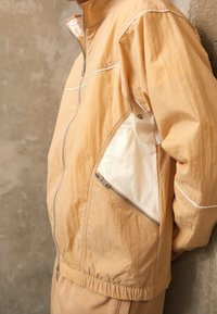 Person som bär en beige och vit zip-jacka med dragkedjefickor, stående mot en strukturerad betongvägg.