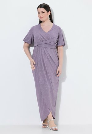 Kobieta ubrana w błyszczącą lawendową sukienkę maxi w stylu kopertowym z krótkimi rękawami i srebrne sandały na wysokim obcasie, stojąca na białym tle.