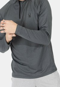 Dunkelgrauer langärmeliger Pullover mit einem Viertelreißverschluss, der ein dezentes Punktemuster und ein Logo auf der Brust aufweist, aus weichem, dehnbarem Material gefertigt.