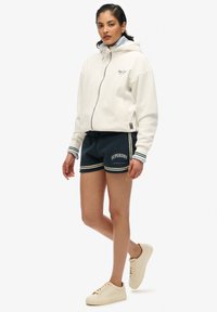 Witte hoodie met ritssluiting, marineblauwe shorts met lichte accenten en tekst, gecombineerd met beige sneakers. Schone en casual uitstraling.