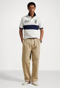 Λευκό πόλο μπλουζάκι με ναυτικό οριζόντιο ρίγες, κοντά μανίκια και γιακά. Μπεζ άνετο παντελόνι, λευκά αθλητικά παπούτσια και ρολόι.