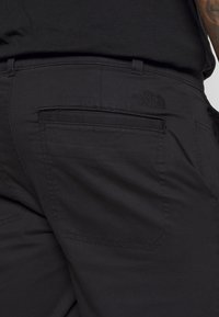 Svarta tygbyxor med en bakficka som har en broderad logotyp. Texturen verkar slät med en skräddarsydd passform.