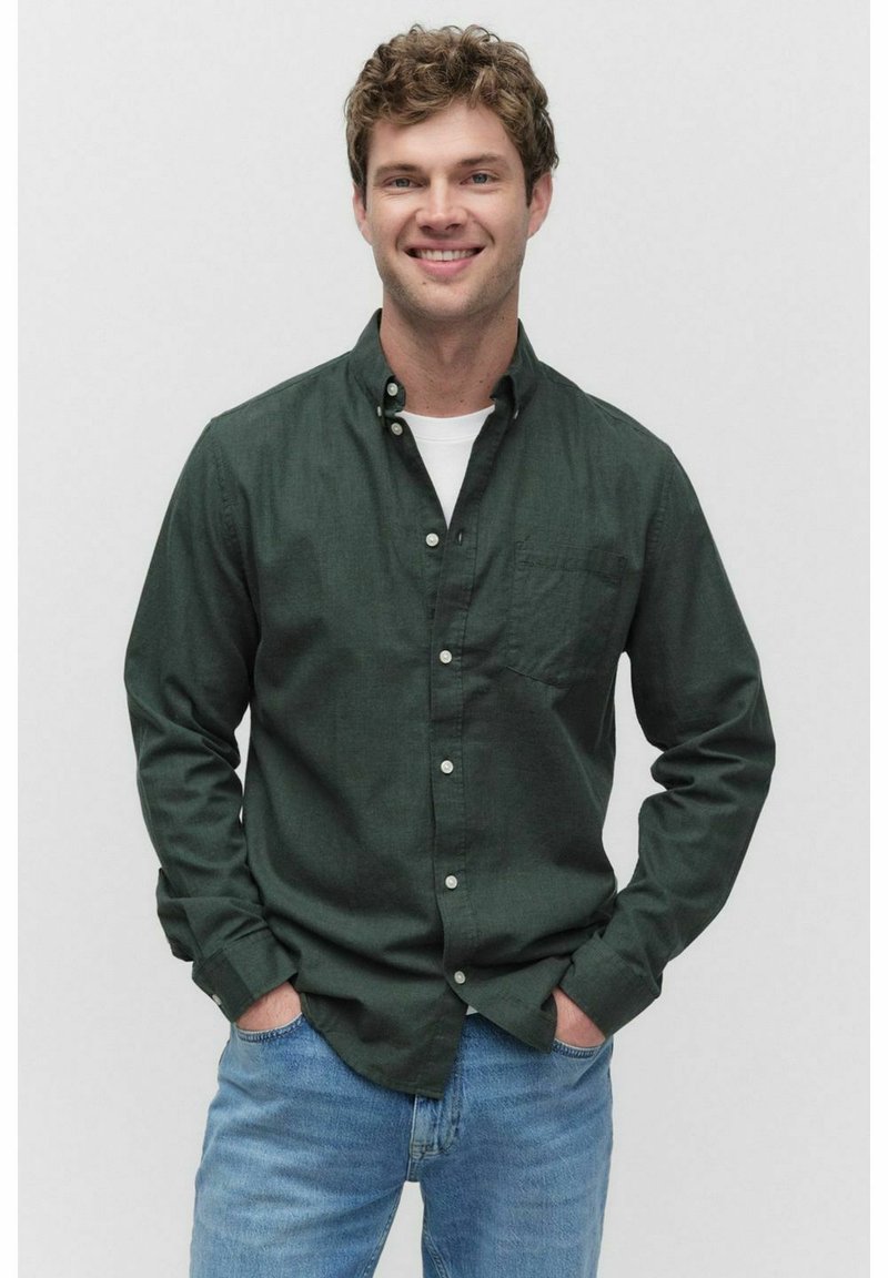 Glunderende man met krullend haar, gekleed in een donkergroen overhemd over een wit T-shirt en lichtblauwe jeans, met zijn handen in zijn zakken tegen een effen achtergrond.