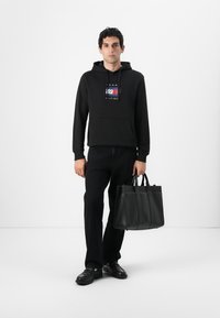 Sort bomulds-hættetrøje med et logo på forsiden, parret med sorte jeans. Modellen holder en struktureret sort læder tote-taske med flere rum.
