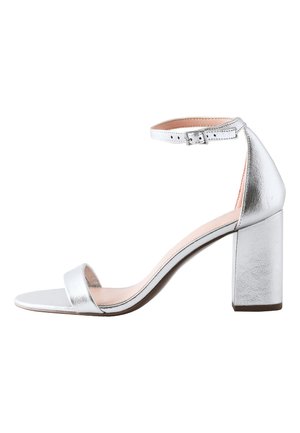 Kvinde i navy ærmeløs kjole og sølv hæle sidder på en kromstol, hviler albuerne på knæene og folder hænderne nær sit ansigt.