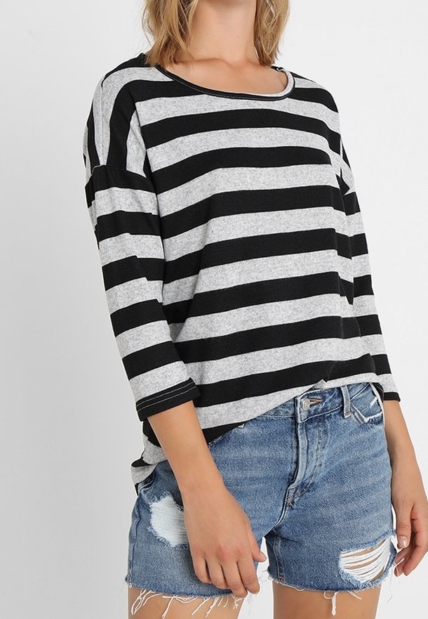 Femme portant un t-shirt à manches longues rayé noir et gris et un short en jean bleu délavé, debout devant un fond blanc uni.
