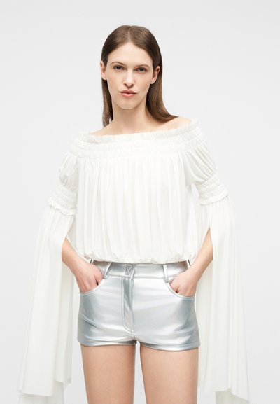 Γυναίκα με ίσια καστανά μαλλιά φοράει λευκή μπλούζα με ακάλυπτους ώμους και μακριά μανίκια, καθώς και μεταλλικά ασημί σορτς με ψηλή μέση, με τα χέρια στις τσέπες.