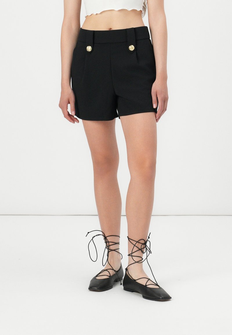 Schwarze maßgeschneiderte Shorts mit zwei vorderen Taschen und Akzenten aus goldenen Knöpfen. Getragen mit schwarzen Schnürflats, die ein spitz zulaufendes Design aufweisen.