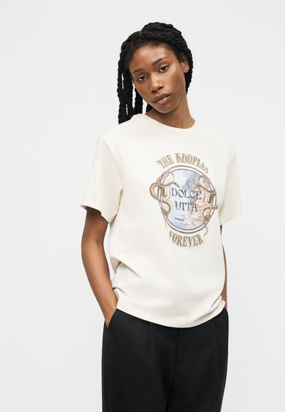 Γυναίκα με κοτσίδα, φορώντας κρεμ γραφικό t-shirt με κείμενο και μαύρο παντελόνι, στέκεται με τα χέρια στην τσέπη μπροστά σε απλό φόντο.