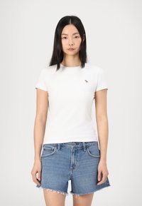 Hvit T-skjorte med kort erm og en liten brun hestelogo, sammen med blå denimshorts med frynsete hem og knappestolpe.