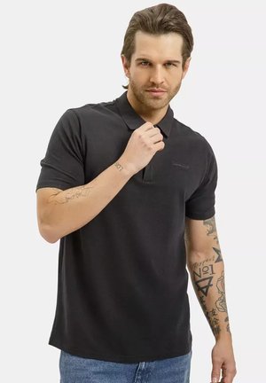 Młody mężczyzna z zaczesanymi do tyłu włosami, ubrany w czarną koszulkę polo i niebieskie dżinsy, pokazuje tatuaże na obu ramionach, pozując z dłonią przy kołnierzu.