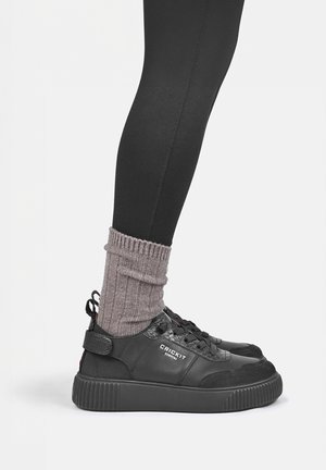 Zijaanzicht van een zwarte sneaker met veters en een riem, gedragen met grijze geribde sokken en zwarte legging op een witte achtergrond.