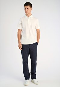 Biała koszulka polo z krótkim rękawem i pionowym prążkowaniem, w zestawieniu z ciemnogranatowymi spodniami i białymi sneakersami, stojąca na neutralnym tle.