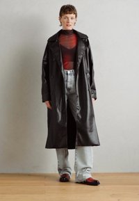 Cappotto lungo in pelle nera con ampi revers, design doppiopetto, indossato sopra un top trasparente rosso e jeans azzurri chiari.
