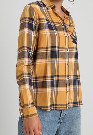 Kobieta ubrana w koszulę na guziki z długim rękawem w żółto-granatowo-brązową kratę, w połączeniu z jasnoniebieskimi dżinsami, na neutralnym tle.