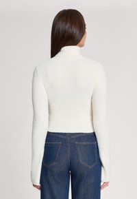 Maglione bianco lavorato a maglia, a maniche lunghe e collo alto, aderente in vita. Abbinato a jeans in denim blu scuro a vita alta.