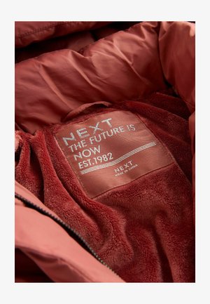 Maroon jacketin sisäosa on pehmeää, tekstuuriltaan fliisiä, jossa on etiketti, joka ilmoittaa "NEXT KATSOMME TULEVAISUUTEEN PERUSTELTU 1982." Vetoketjullinen avaus ylhäällä.