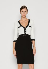 Robe noir et blanc avec des manches trois-quarts, décolleté en V et détails en boutons. Présente une bordure contrastante et une ceinture décorative à la taille.