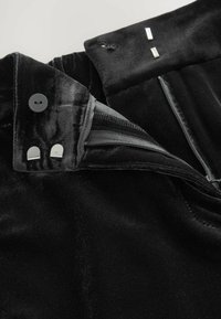 Indumento in velluto nero con apertura a zip e chiusura a bottoni; presenta dettagli in hardware argentato e una texture liscia. Vista ravvicinata.