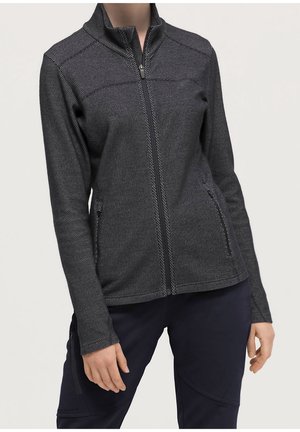 Vrouw draagt een aansluitend zwarte en grijze gestructureerde jas met ritssluiting en ritssluitzakken aan de zijkanten en een donkere broek tegen een effen achtergrond.