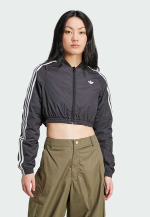 Veste noire courte avec fermeture éclair à l'avant, poignets côtelés et rayures blanches sur les côtés. Présente un petit logo blanc sur la poitrine gauche. Associée à un pantalon vert olive.